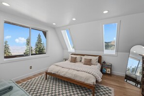 A bedroom with a large bed, two windows, a skylight, and a view of trees and mountains.