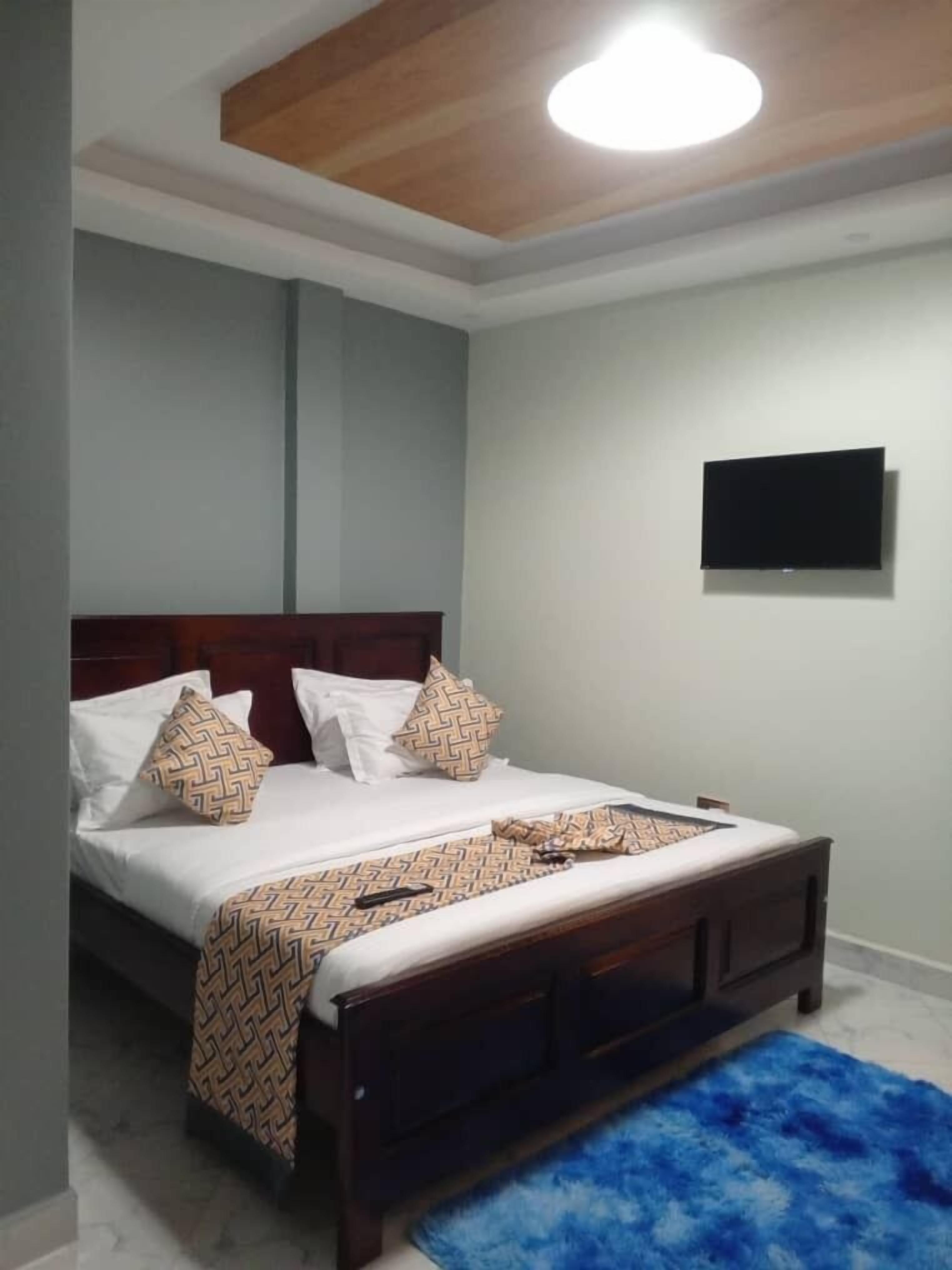 A bedroom with a bed, a wooden headboard, a blue rug, and a mounted TV on the wall.
