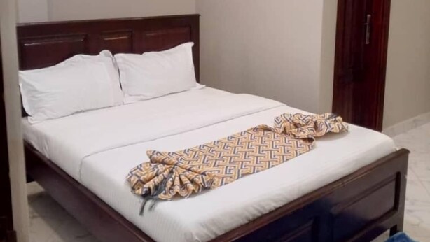 A bedroom with a bed, a wooden headboard, a patterned throw blanket, and a blue rug. - AYEBA ZION HOTEL (Fort Portal)