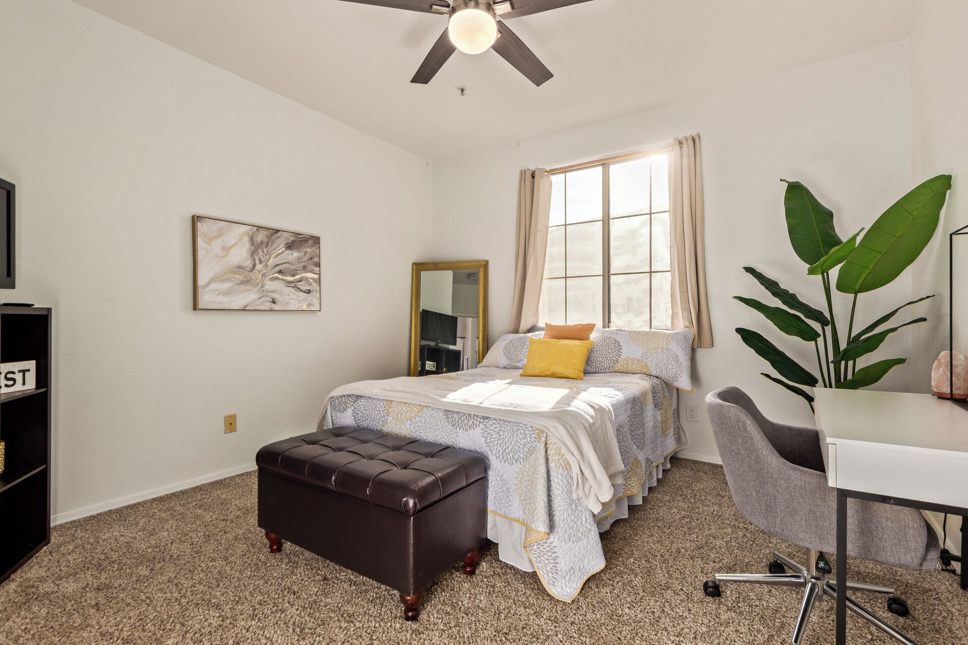 A neatly arranged bedroom with a bed, a desk, a chair, a large plant, and a ceiling fan.