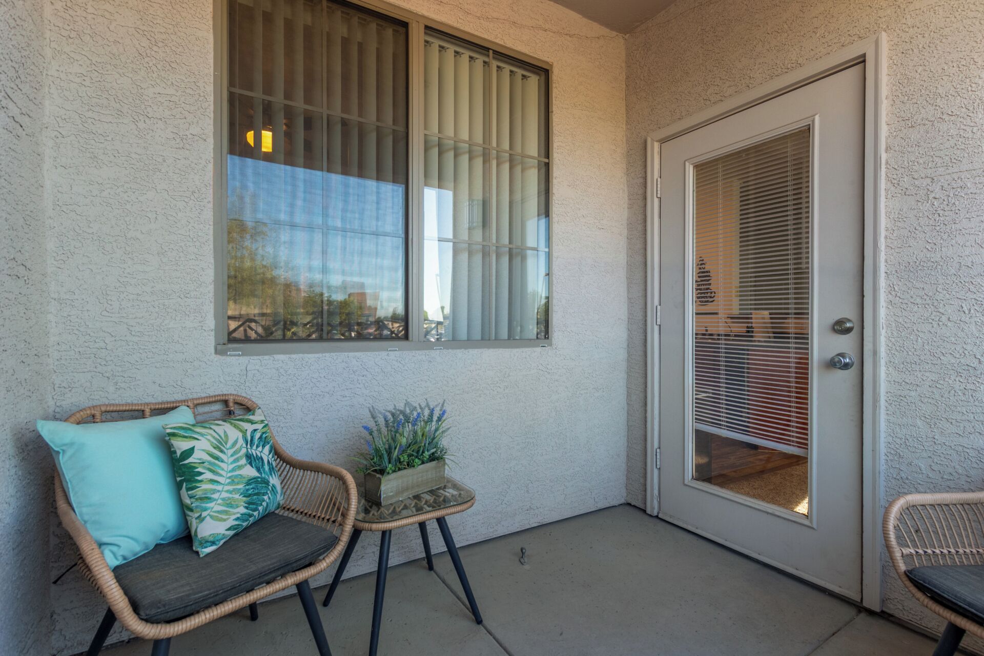 A small porch with a wicker chair, a small table with a plant, and a door with a window.