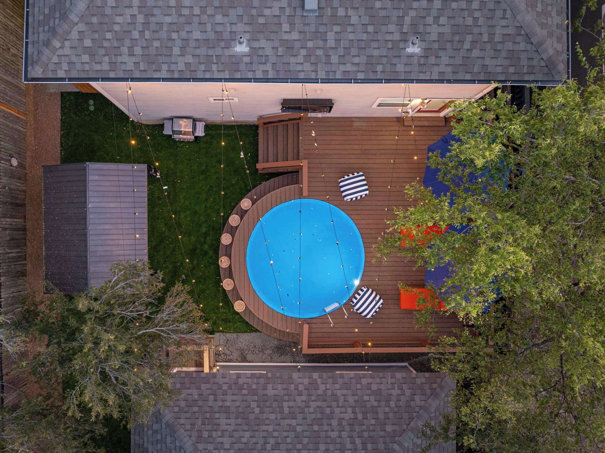 An aerial view of a backyard with a circular pool, a wooden deck, and surrounding greenery.