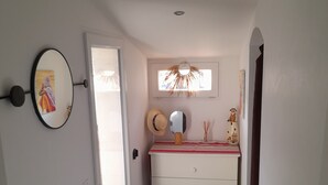 Um corredor estreito com uma porta branca, um espelho, uma pequena mesa com um chapéu e um enfeite na parede.