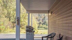 A covered outdoor area with a grill and chairs, a garage door, and a view of trees and a road.