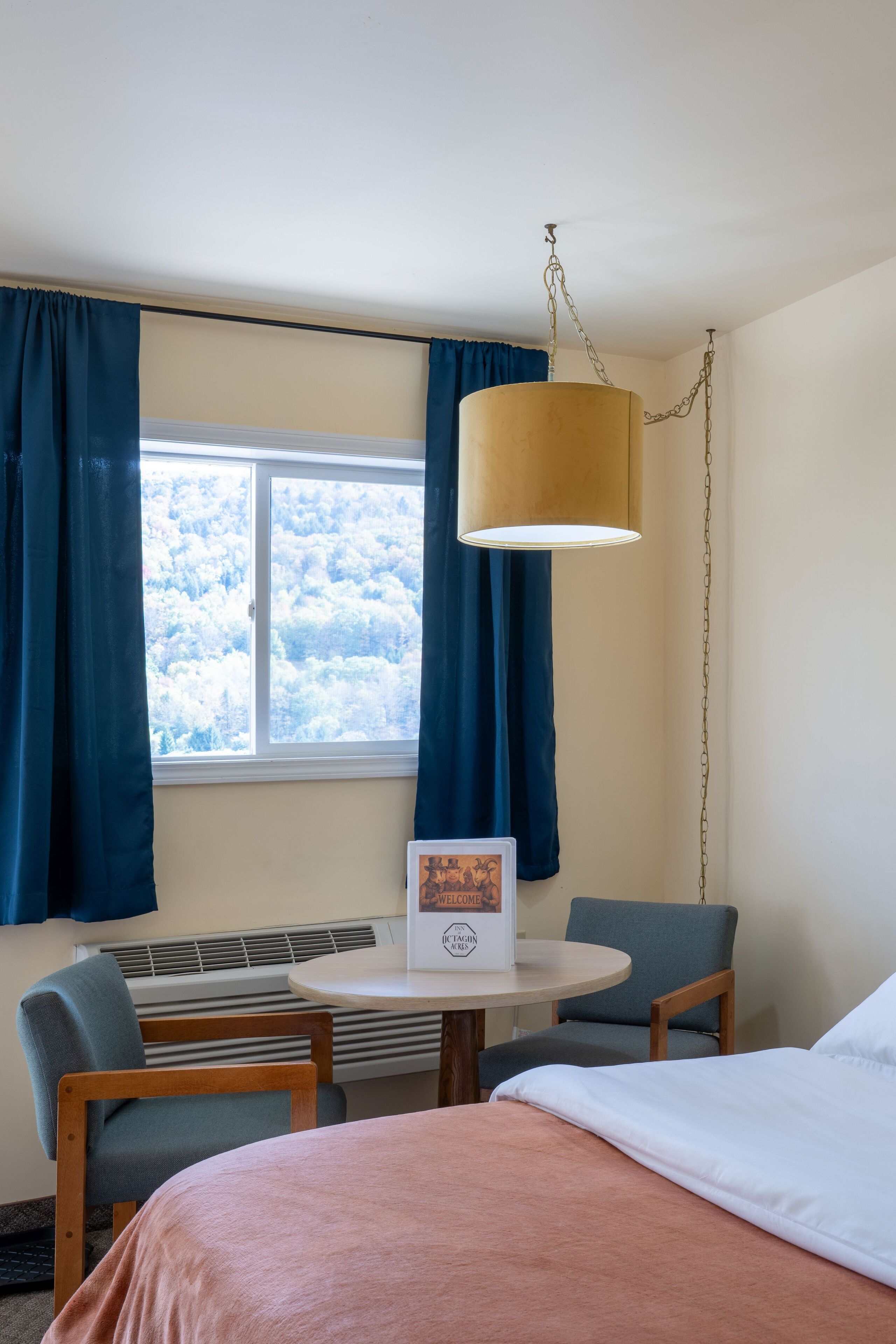 Hotellihuone, jossa on sänky, kaksi tuolia, pieni pöytä ja ikkuna, jossa on verhot.