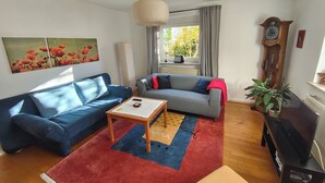 A living room with a blue sofa, a grey sofa, a coffee table, a red rug, a wooden floor, a large window with curtains, a tall wooden clock, and a potted plant. - Vacation apartment in the countryside near Ulm in the "small Lautertal" - with garden (Blaustein)