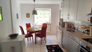 A compact kitchen with white cabinets, a dining table with chairs, a black and red patterned rug, and a window providing natural light. - Vacation apartment in the countryside near Ulm in the "small Lautertal" - with garden (Blaustein)