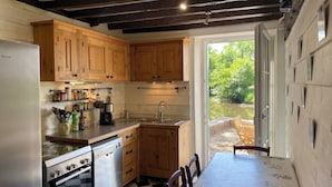 A kitchen with wooden cabinets, a dishwasher, a stove, and a sink. There is a table with chairs and a view of a garden through the open door. - Mill on the banks of the Indre in Berry. A bucolic, historic site on an island (Ardentes)