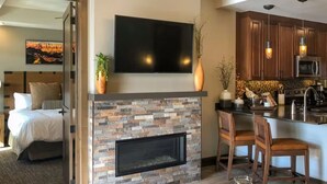 A modern living room with a fireplace, a TV mounted above it, and a kitchen area with wooden cabinets and pendant lights.