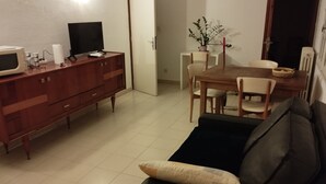 A living room with a dark sofa, a wooden dining table with chairs, a TV on a wooden cabinet, and a potted plant on the table.