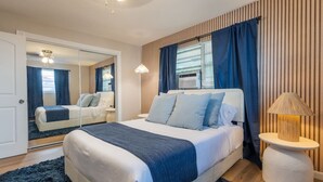 A modern bedroom with a large bed, a bedside table with a lamp, a mirror, and a window with curtains. - Comfort & Style Modern | 2BR In West Palm Beach (West Palm Beach)