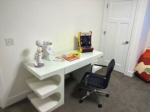 Ein weißer Schreibtisch mit einem schwarzen Bürostuhl, einem Pac-Man-Automaten und zwei Astronautenfiguren.