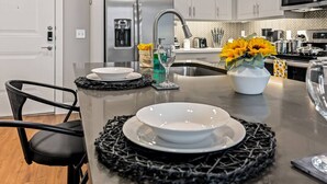 A modern kitchen with a stainless steel refrigerator, a sink, and a microwave. There are two black chairs and a dining table with white plates and a vase of yellow flowers. - Cozy Luxury 1bd | Pool+Gym+Stockyards+Free Parking (Fort Worth)