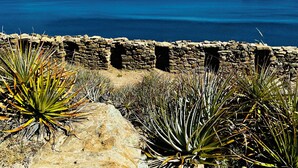 Un paysage côtier avec un mur de pierre et des plantes d’agave au premier plan, qui s’ouvre sur une vue de l’océan et de montagnes lointaines.