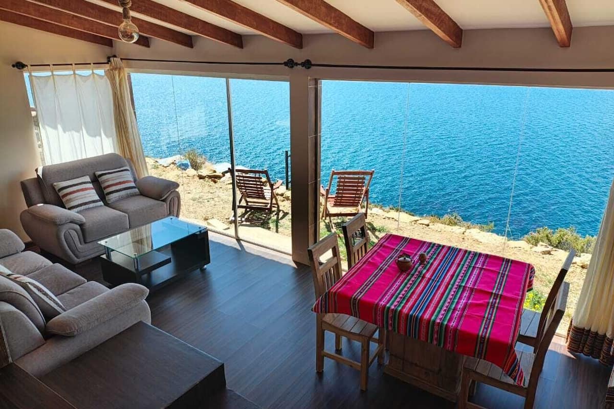 Un vaste espace de vie comprenant un ensemble de canapés, une table basse en verre et une table à manger avec des chaises. La pièce est dotée de grandes fenêtres offrant une vue sur la mer.