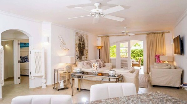 A spacious living room with a white sofa, a coffee table, and a ceiling fan. There is a view of greenery outside through large windows. - Sugar Breeze #b106 - by Zenbreak (Holetown)