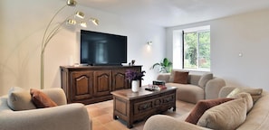 A living room with a large flat-screen TV, a wooden cabinet, a coffee table with decorative items, a sofa, and a floor lamp.