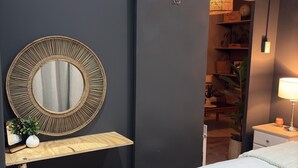 Une chambre à coucher comprenant un lit, une table de chevet avec une lampe, un miroir, un petit tabouret, un panier et une armoire.