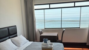 A hotel room with a bed, a small table, a view of the ocean, and a window with curtains. - Los Esteros (Huanchaco)