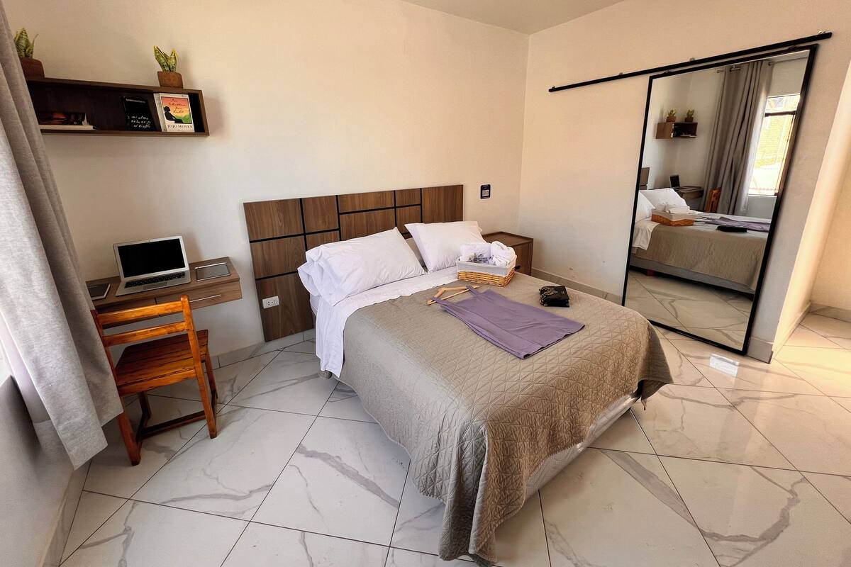 A hotel room with a bed, a desk with a laptop, a mirror, and a shelf with books and plants.