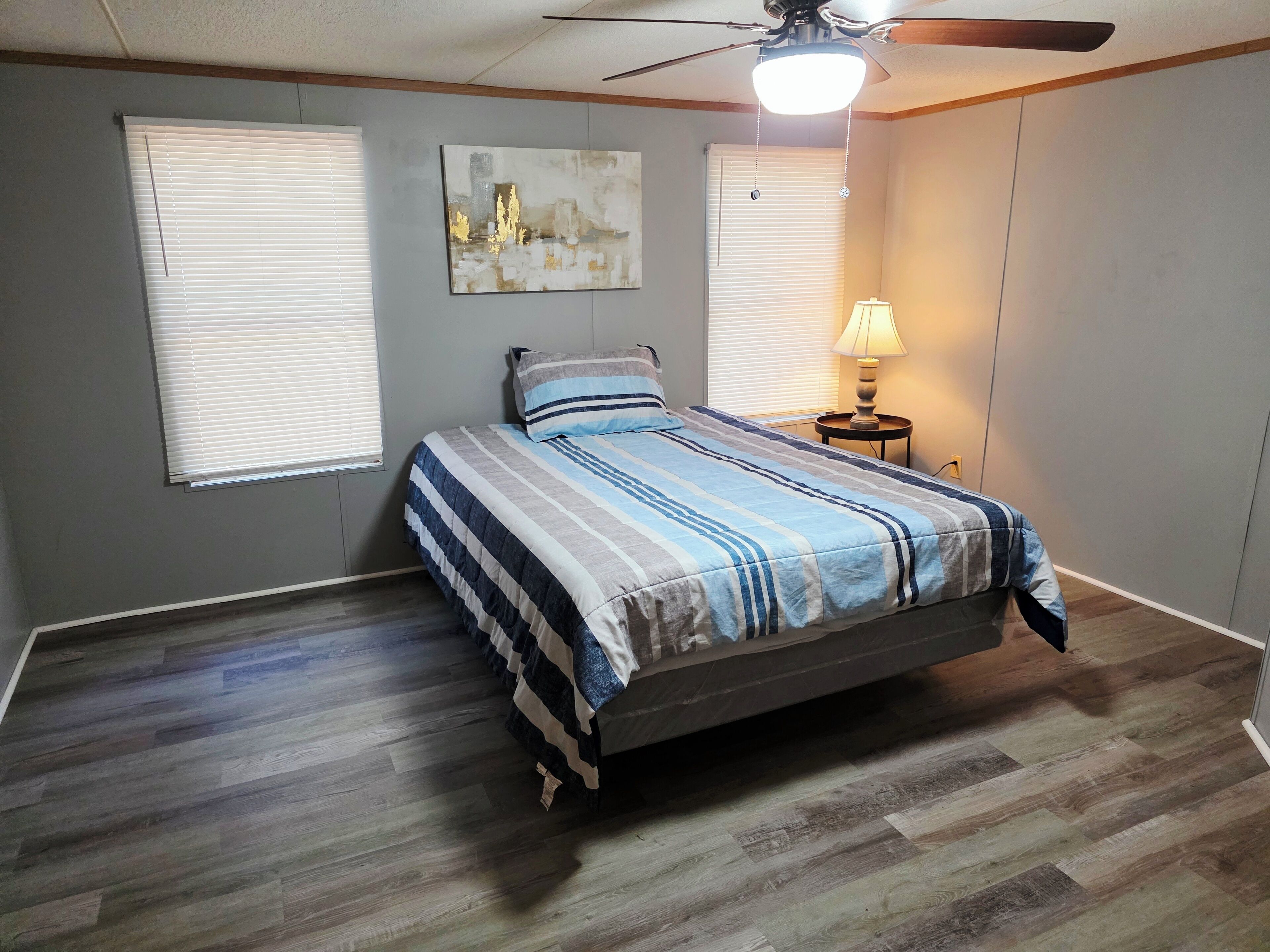 A bedroom with a bed, two windows, a ceiling fan, and a nightstand with a lamp.