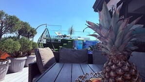 Een terras met een houten tafel, een rieten mand met ananassen en perziken, en een hangstoel.