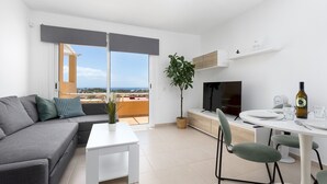 Un salon moderne avec un canapé gris, une table basse blanche, un coin repas avec une table et des chaises blanches, un téléviseur et un balcon avec vue.