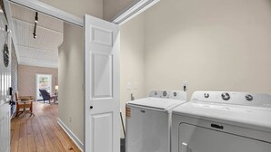 Un lavadero con dos lavadoras y secadoras de carga frontal, piso de madera y una puerta que da a una habitación con pared de ladrillo y una silla.