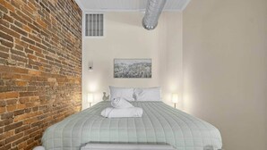 Habitación de hotel con pared de ladrillo, cama con colcha acolchada, dos lámparas de noche y un cuadro enmarcado en la pared.