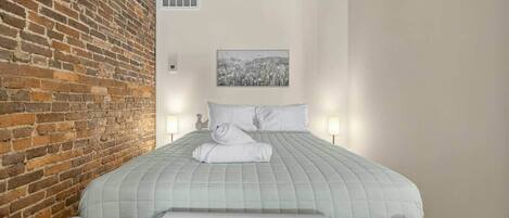 A hotel room with a brick wall, a bed with a quilted bedspread, two bedside lamps, and a framed picture on the wall.