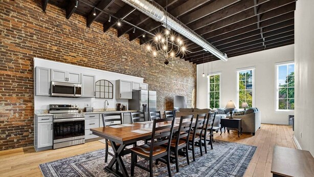 A spacious kitchen with modern appliances, a dining area with a large wooden table, and a living room with a sofa and a TV. - Sleeps 6. Two Downtown Lofts Combined. (Fayetteville)