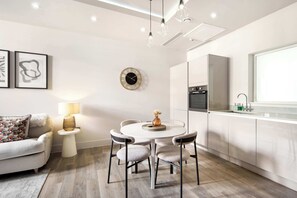 A modern kitchen with white cabinets, a dining table with chairs, a round wall clock, and a sofa with a patterned pillow.