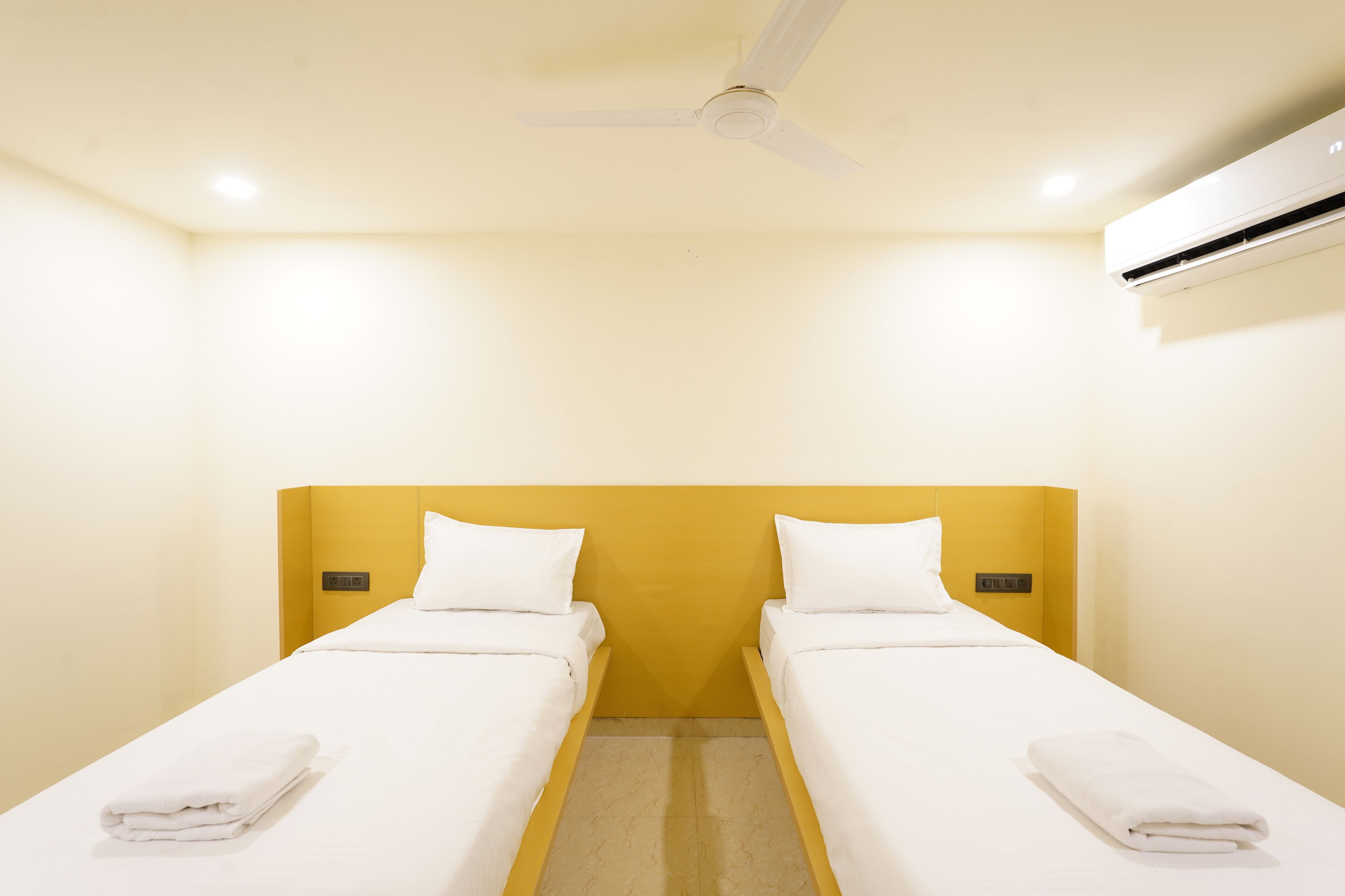 A hotel room with two single beds, a ceiling fan, an air conditioning unit, and a light switch on the wall.