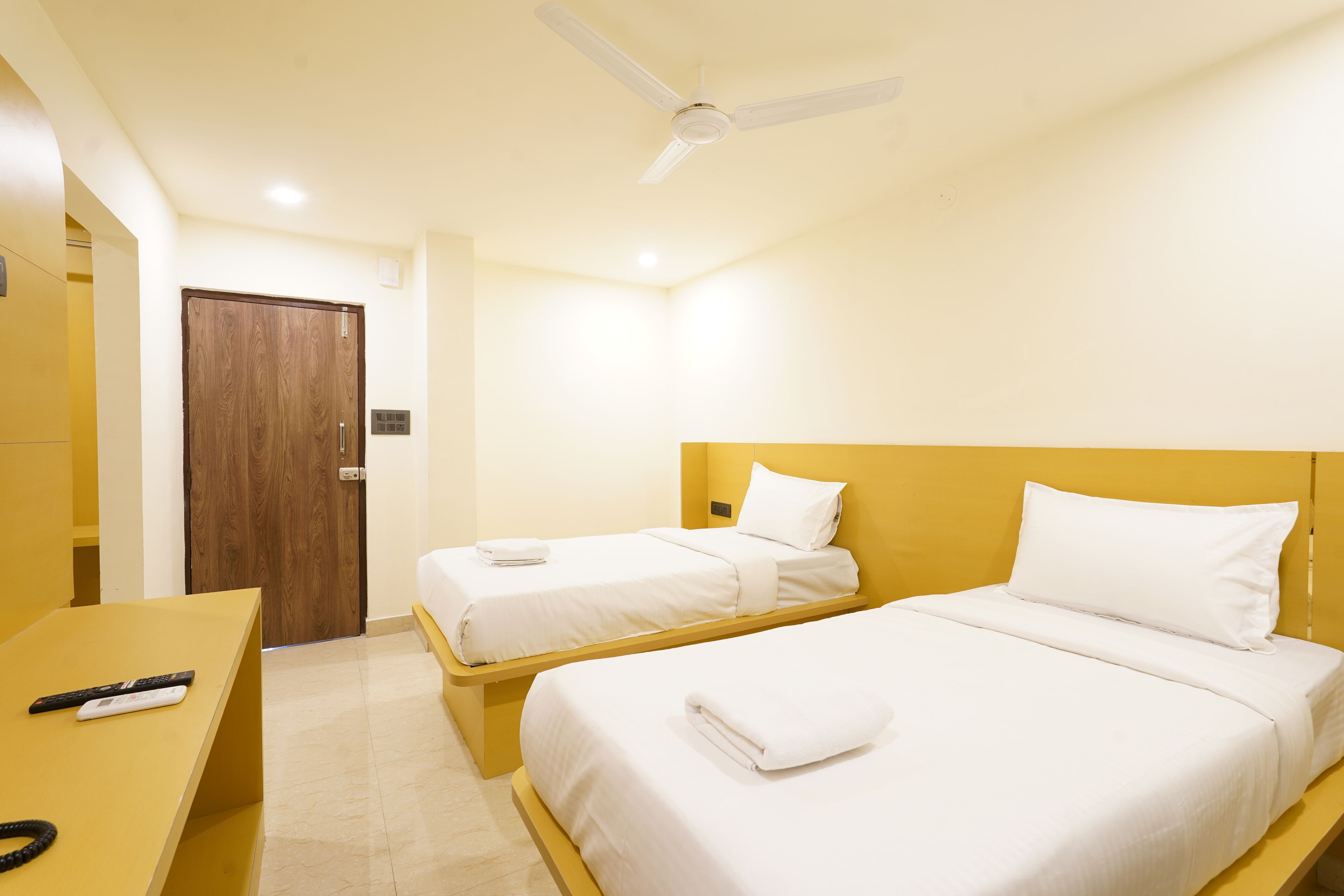A hotel room with two single beds, a wooden door, a desk with a remote control, and a ceiling fan.