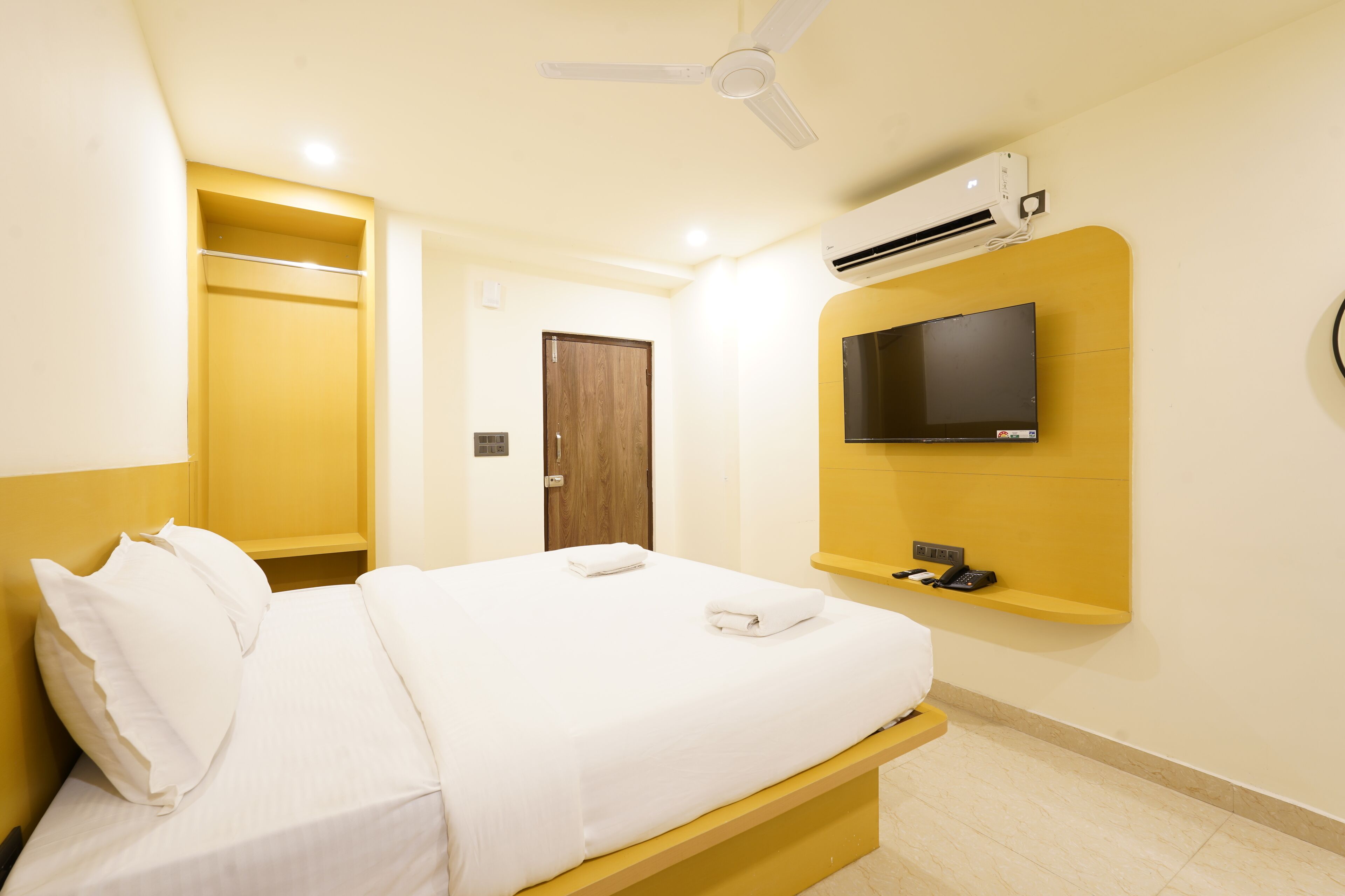 A hotel room with a bed, a TV mounted on the wall, an air conditioning unit, and a ceiling fan.