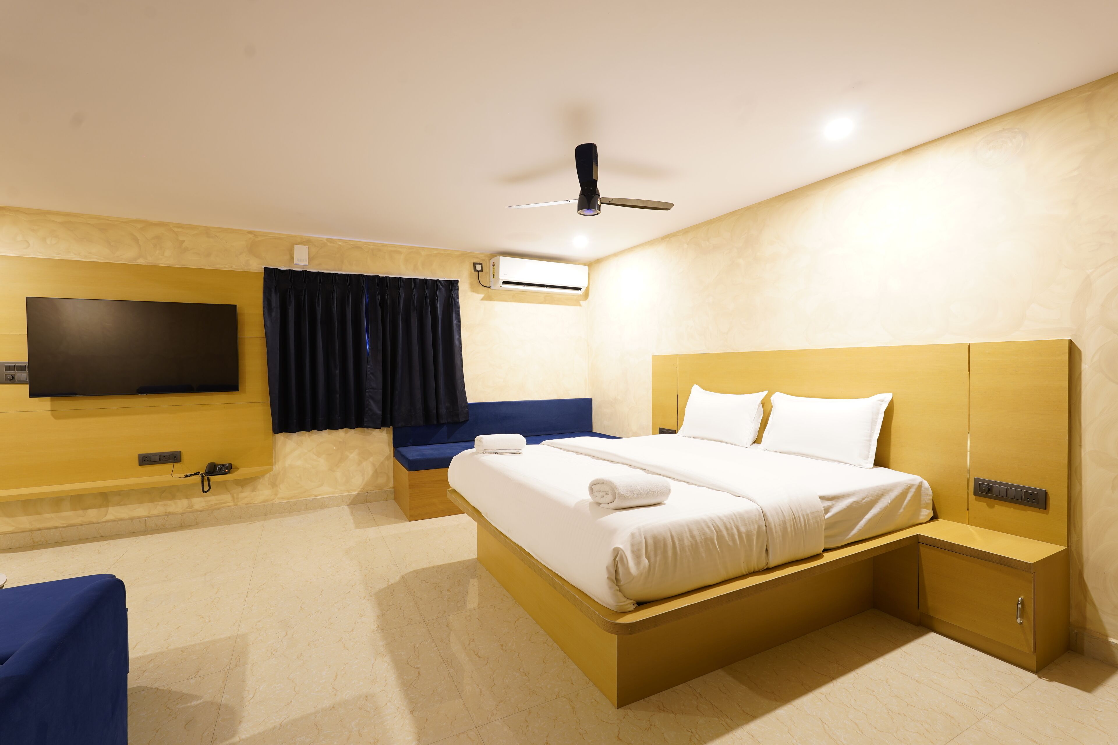 A modern hotel room with a large bed, a flat-screen TV mounted on the wall, a ceiling fan, and an air conditioning unit.