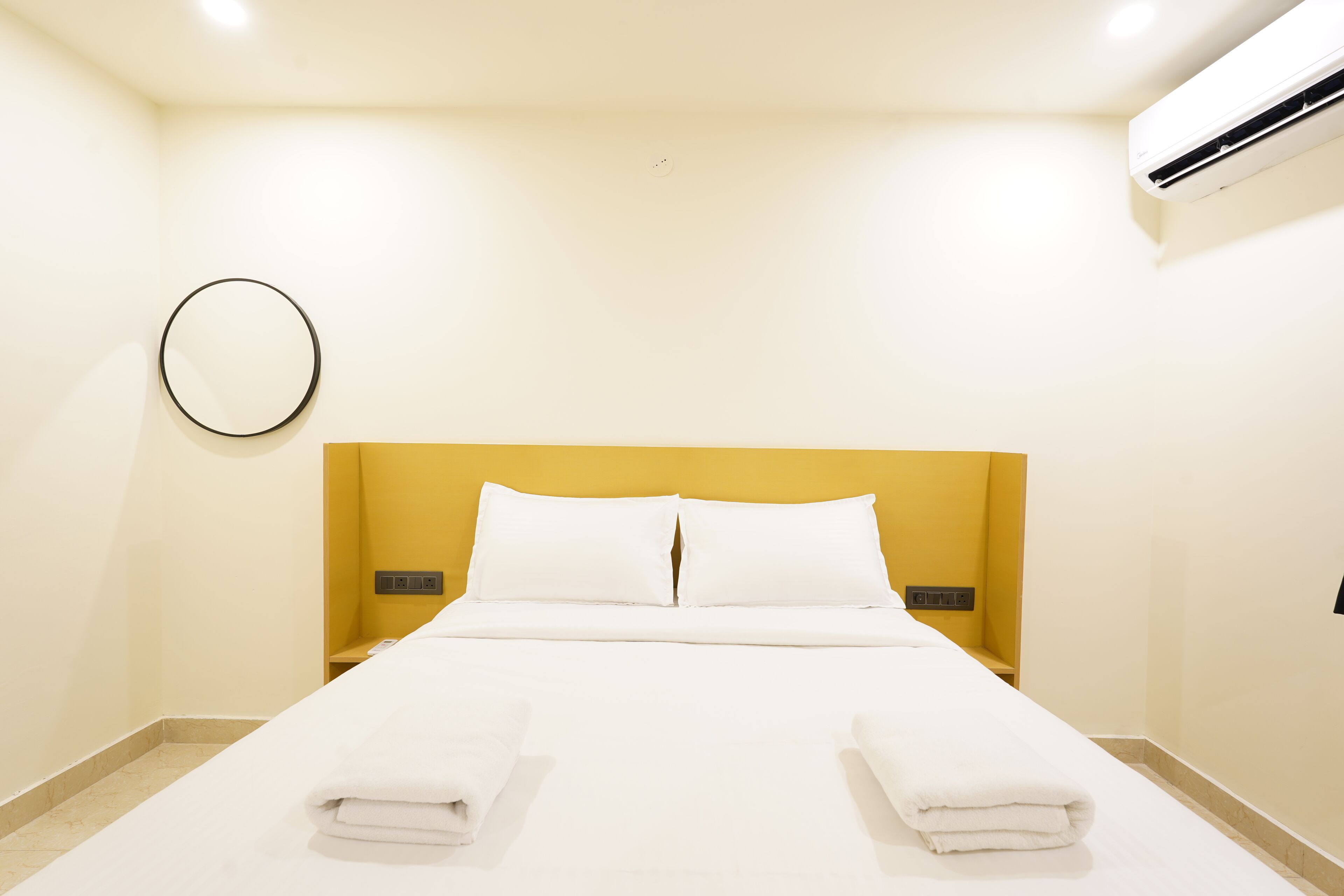 A hotel room with a bed, white linens, a yellow headboard, a round mirror, and an air conditioning unit.