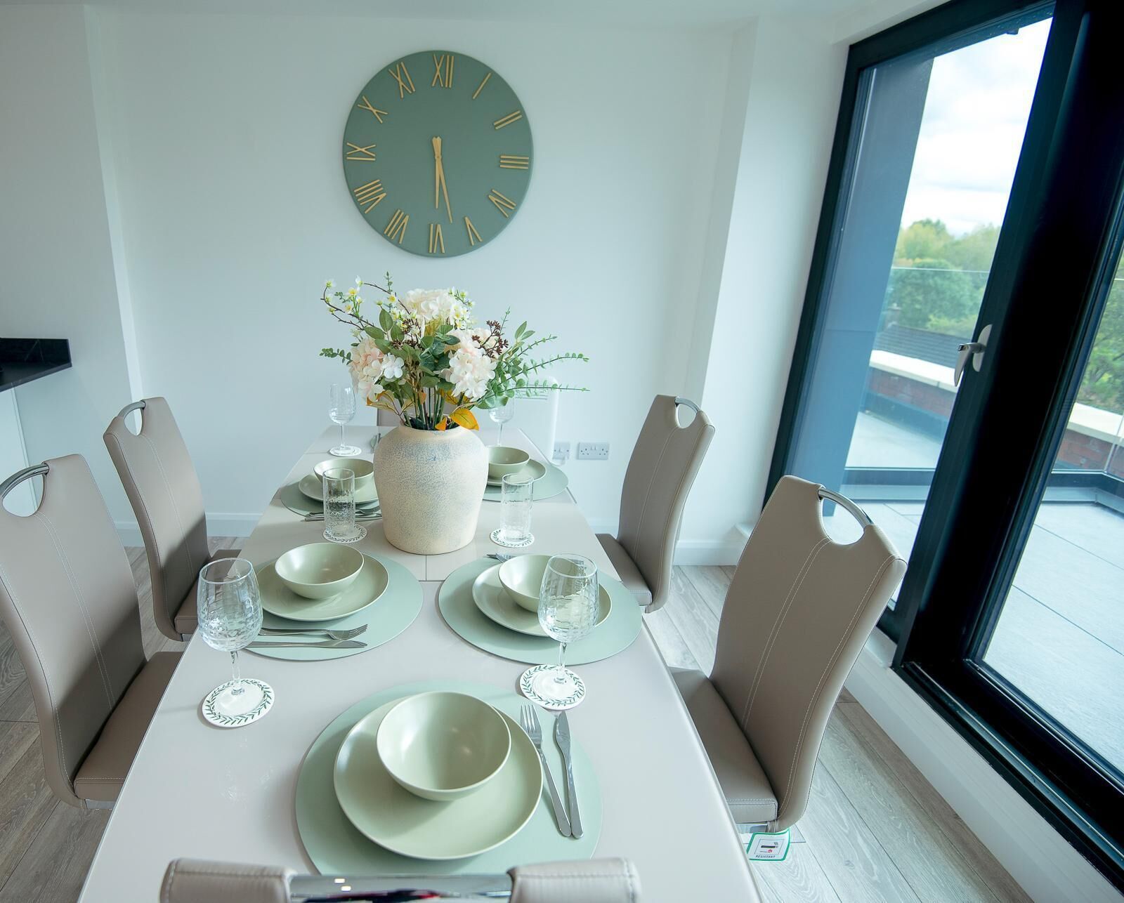 A dining room with a round table set for four, a vase of flowers, and a large wall clock.