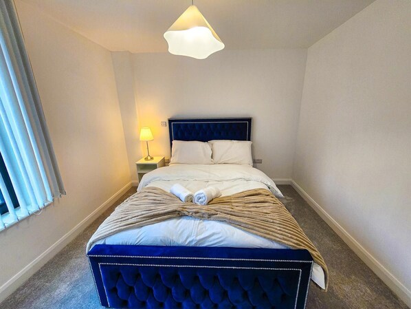 Ein ordentlich bezogenes Bett mit blauem Kopfteil, einer Nachttischlampe und einem Fenster mit Jalousien.