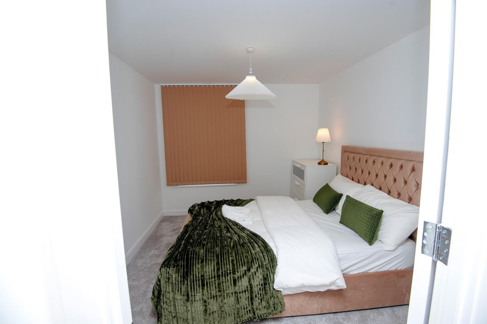 A bedroom with a bed, a nightstand, a lamp, a window with blinds, and a hanging light fixture.