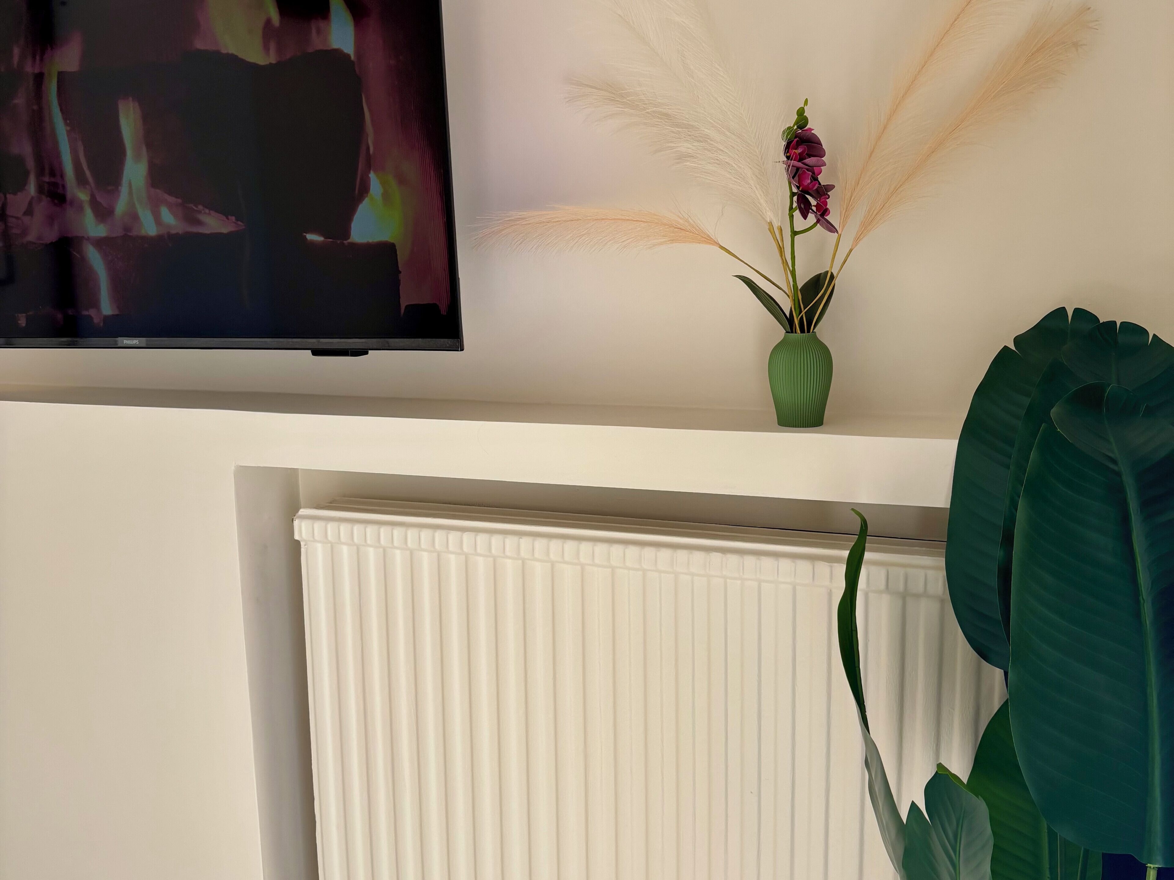 Een woonkamer met een flatscreen-televisie aan de muur, een radiator onder de televisie, en een groene vaas met een plant op een plank.
