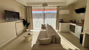 Ein modernes Wohnzimmer mit einer Couch, einem Couchtisch und einem Fernseher. Es gibt eine Küchenzeile mit Schränken und Geräten.