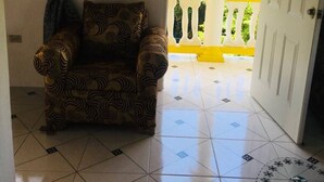 Ein Zimmer mit einem gemusterten Sessel, Fliesenboden, einer weißen Tür und einem Balkon mit gelben Geländern.