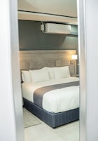 Una cama bien hecha con ropa de cama blanca y un detalle en gris, una lámpara de noche y un aire acondicionado instalado en la pared.