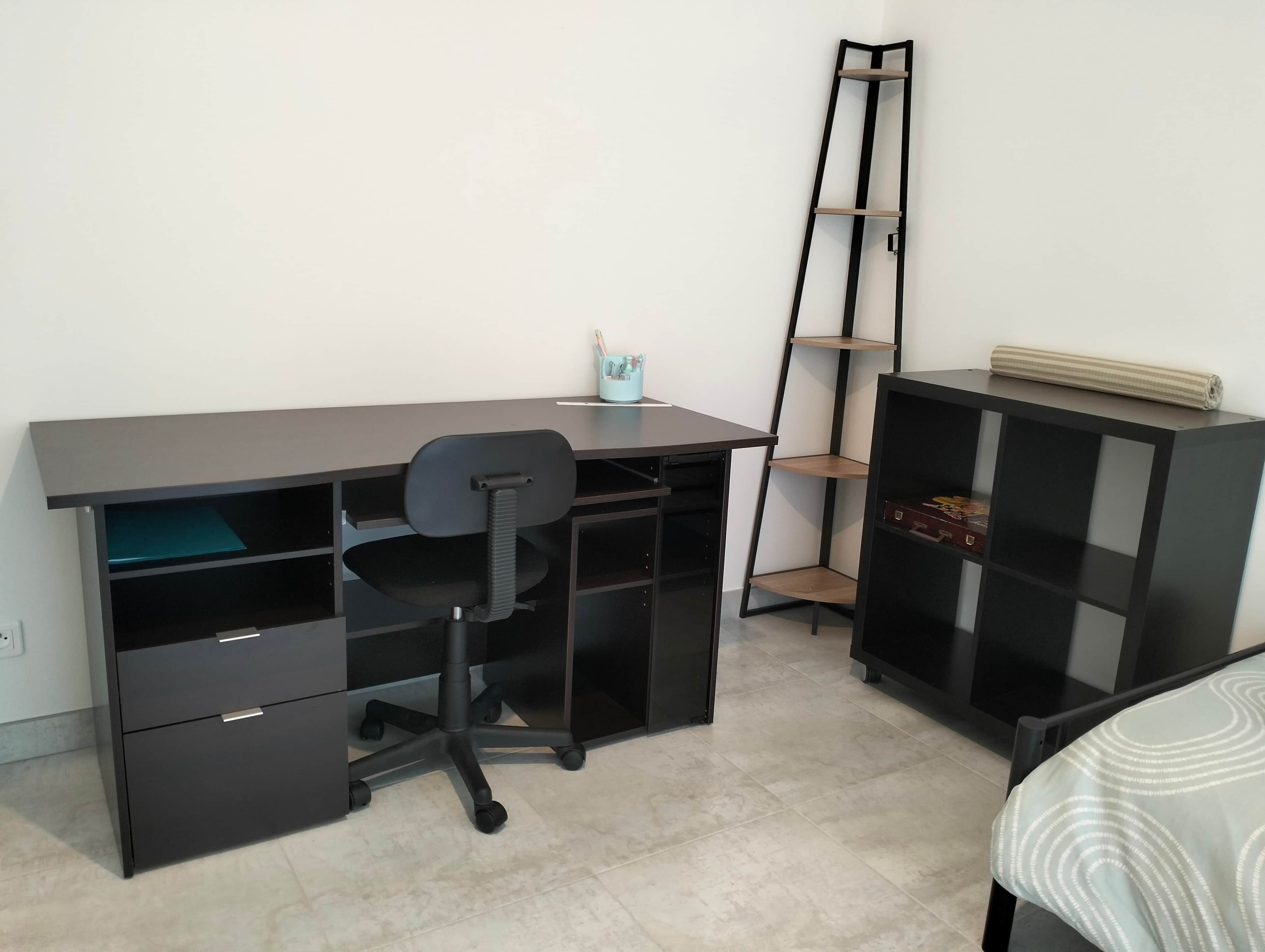 A modern office space with a desk, chair, shelving unit, and a bed.
