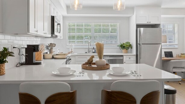 A modern kitchen with white cabinetry, a central island with seating, pendant lighting, and a large window with blinds. - Spacious 2BR prime location | Free Parking & Crib (Tampa)