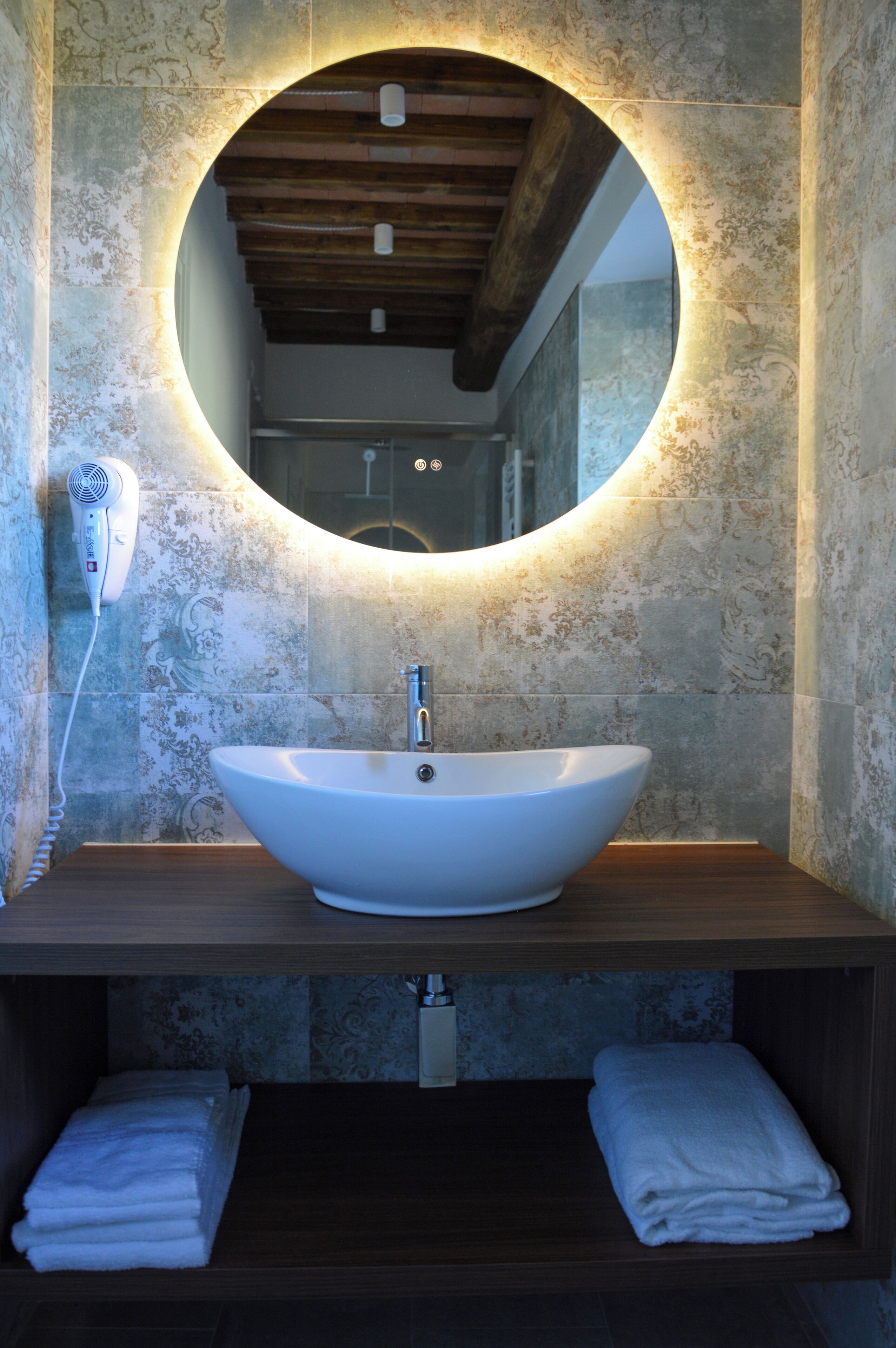 Une salle de bain avec un miroir rond, un lavabo blanc et un sèche-cheveux fixé au mur.