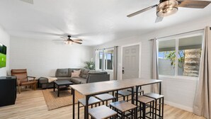 Un vaste espace de vie comprenant un canapé, une table basse et une table à manger avec des chaises. On y trouve également des ventilateurs de plafond, une grande fenêtre avec des rideaux et une porte donnant sur l’extérieur.