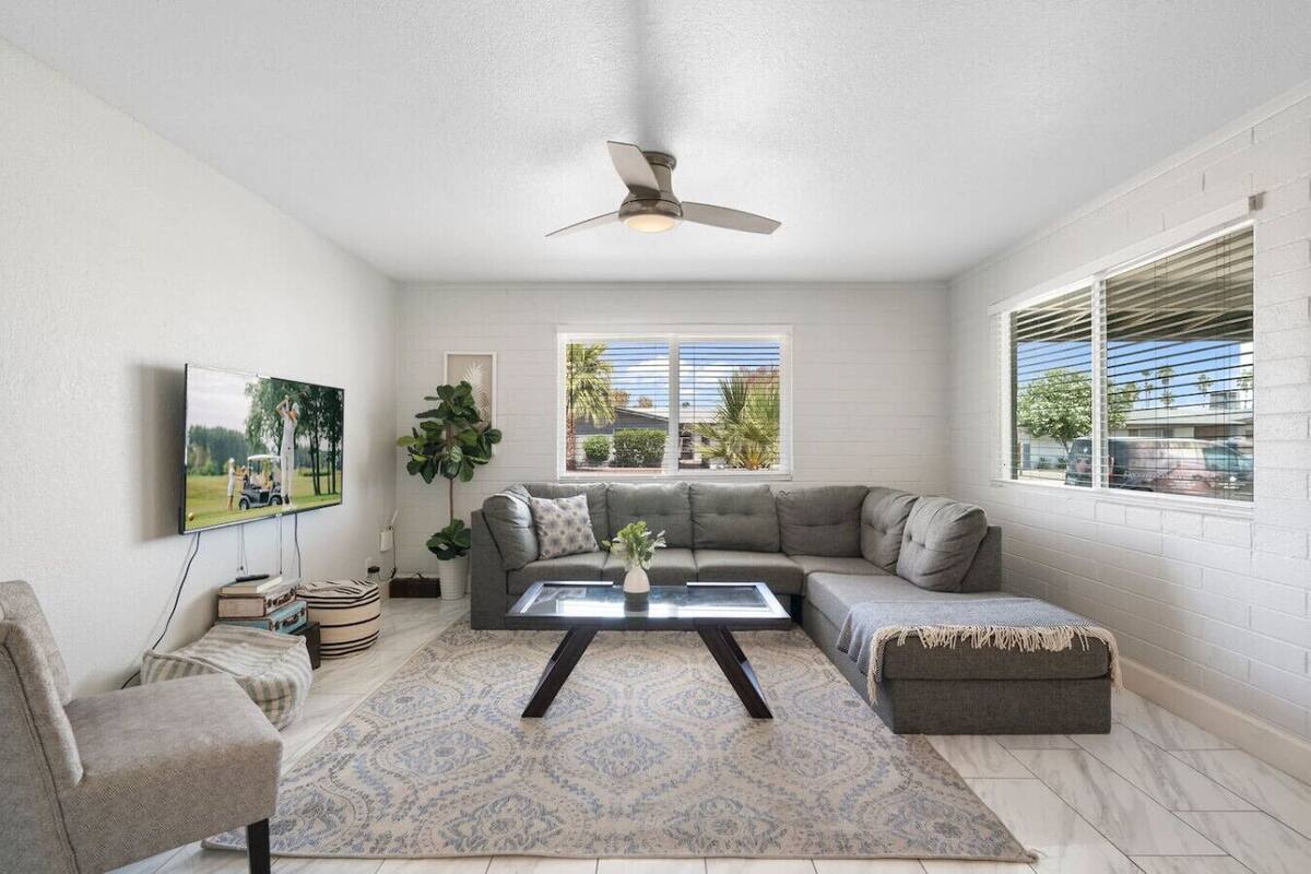 A modern living room with a sectional sofa, a coffee table, a TV mounted on the wall, and large windows with blinds.