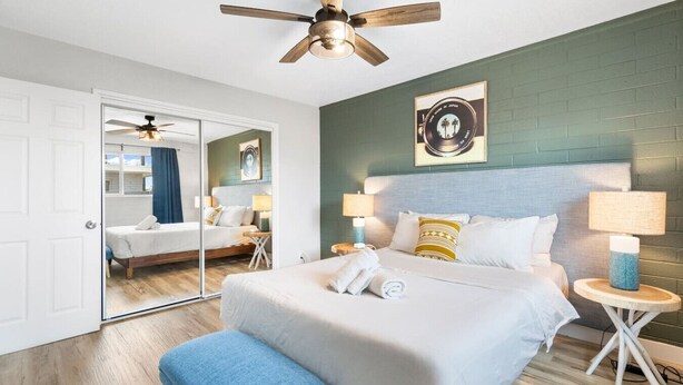 Une chambre d’hôtel avec un grand lit, deux tables de chevet avec des lampes, un ventilateur de plafond, un miroir et une œuvre d’art accrochée au mur.